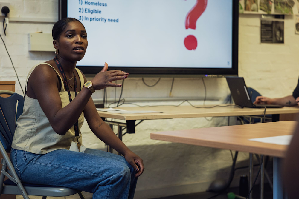 A Shelter colleague sits in one of the hub meeting rooms, with a presentation screen behind her. She is in discussion with teammates who sit off-camera.