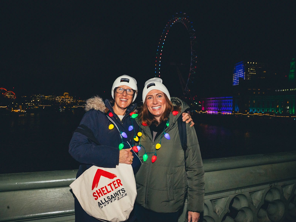 Two Walk for Home participants wrapped in fairy lights, wearing AllSaints x Shelter beanie hats and tote bags.