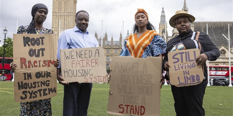 Four Shelter campaigners holding signs on cardboard stand outside Westminster. The signs say statements like: 'root out racism in our housing system' and 'still living in limbo'.