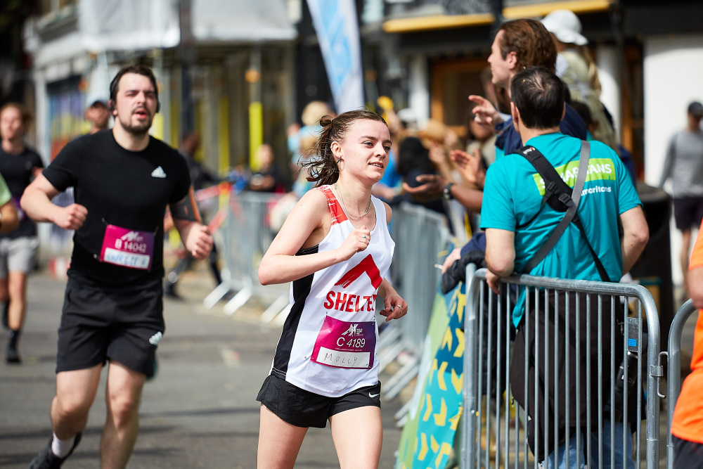 A white woman in a Shelter vest runs past race spectators, looking focused and determined.