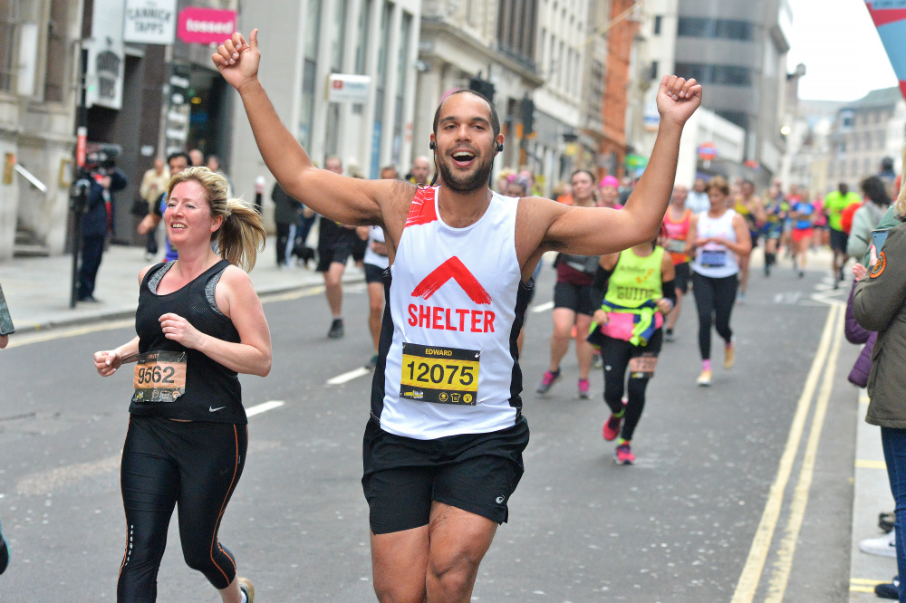 A man wearing a Shelter vest running a city race - he raises his arms in celebration as he passes the cheer point.