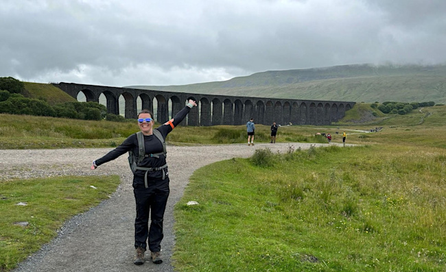 A woman wearing hiking gear holds her arms wide on a pathway, with an impressive view of a viaduct behind her.