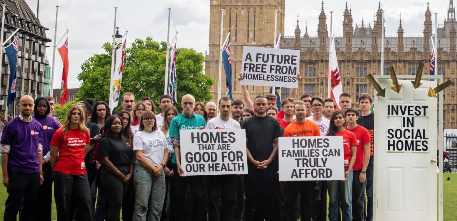 A large group of campaigners standing opposite the Houses of Parliament, holding signs reading 'A future free from homelessness', 'Homes that are good for our health' and 'Homes families can truly afford'. Next to them is a door with the signage 'Invest in social homes'.