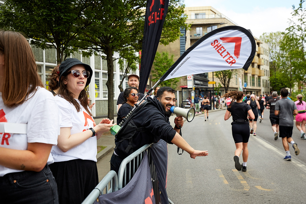 The Shelter Events team at the cheer point, wearing Shelter T-shirts and holding banners. One person holds a megaphone and he reaches out to give runners high-fives.