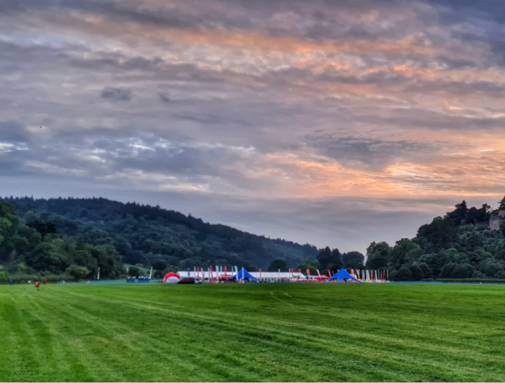 The challenge camp at sunset, with a big lit-up marquee and finish line banners. The camp is surrounded by green fields and trees.