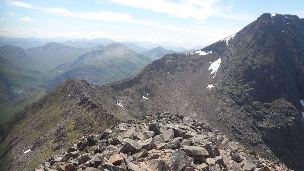 The summit of Ben Nevis, with patches of snow on the peak and a stunning view of other mountains in the background.
