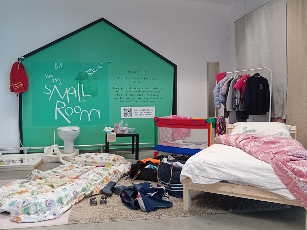 An installation of a child's room living in temporary accommodation is installed in an IKEA showroom. There is a toilet, clothes in a suitcase, a bed, and a sleeping bag on the floor. There is a quote from a child talking about living with mice shown on a house sign.