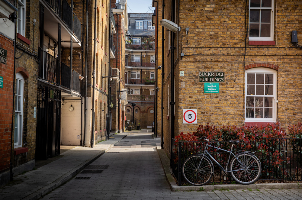 A block of social housing flats visible at the end of an alleyway, with colourful flowers on the balconies. A bicycle leans against the railings at the front of the building.