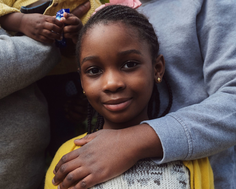 A child wearing a yellow jumper looks into the camera smiling, with her big sister's arm protectively around her.