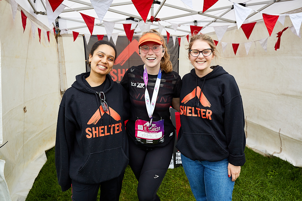 Two female Events team members wearing black Shelter hoodies stand either side of a woman with a medal.