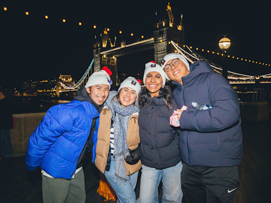 A group of four people wearing Walk for Home white and red bobble hats pose for a photo. It's night time and the lights of Tower Bridge are in the background.