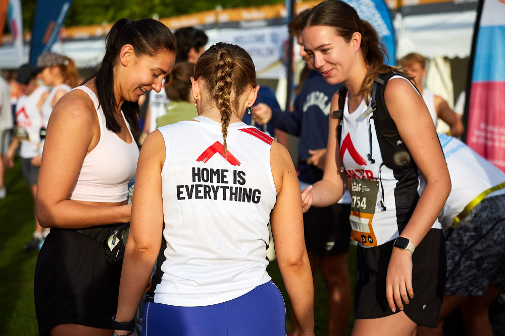 Three women runners wearing Shelter vests stand together chatting before the race starts. The back of their vests read 'Home is everything'.