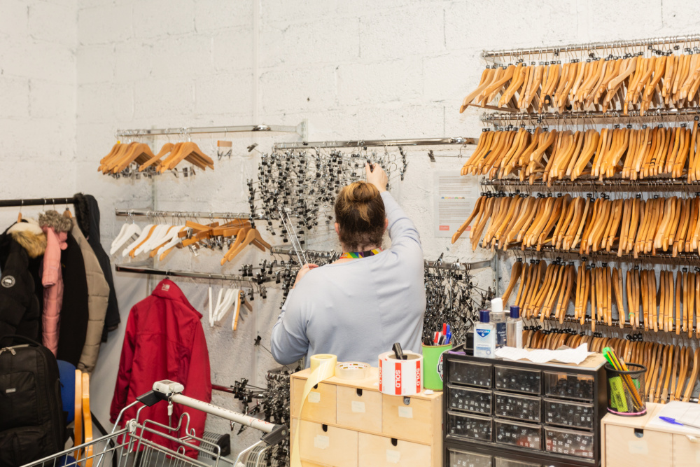 Behind the scenes at a Shelter shop, a woman volunteer selects clothes hangers from a wall filled with rails of them.