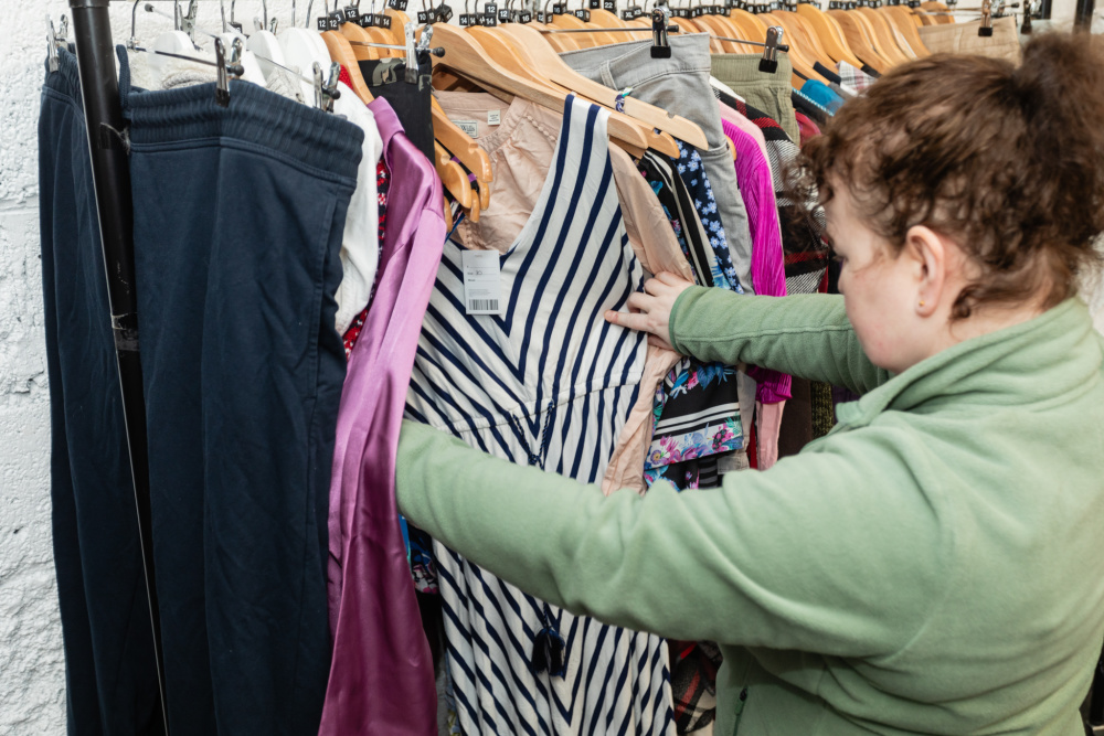 A white woman with brown hair in a ponytail looks through a clothes rail in a Shelter shop. She has pulled out a V-neck dress with blue and white stripes to take a closer look.