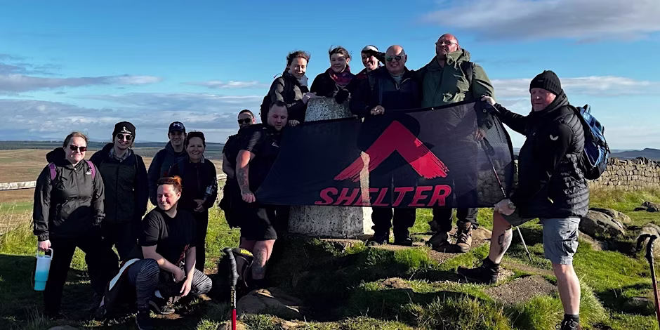 A group of hikers on a sunny day, standing on Hadrian's Wall and holding up a Shelter banner.