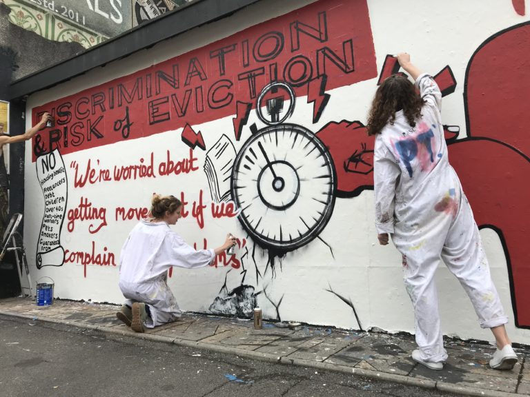 Two Bristol campaigners painting the mural, with a ticking clock illustration next to the title 'Discrimination & Risk of Eviction'.