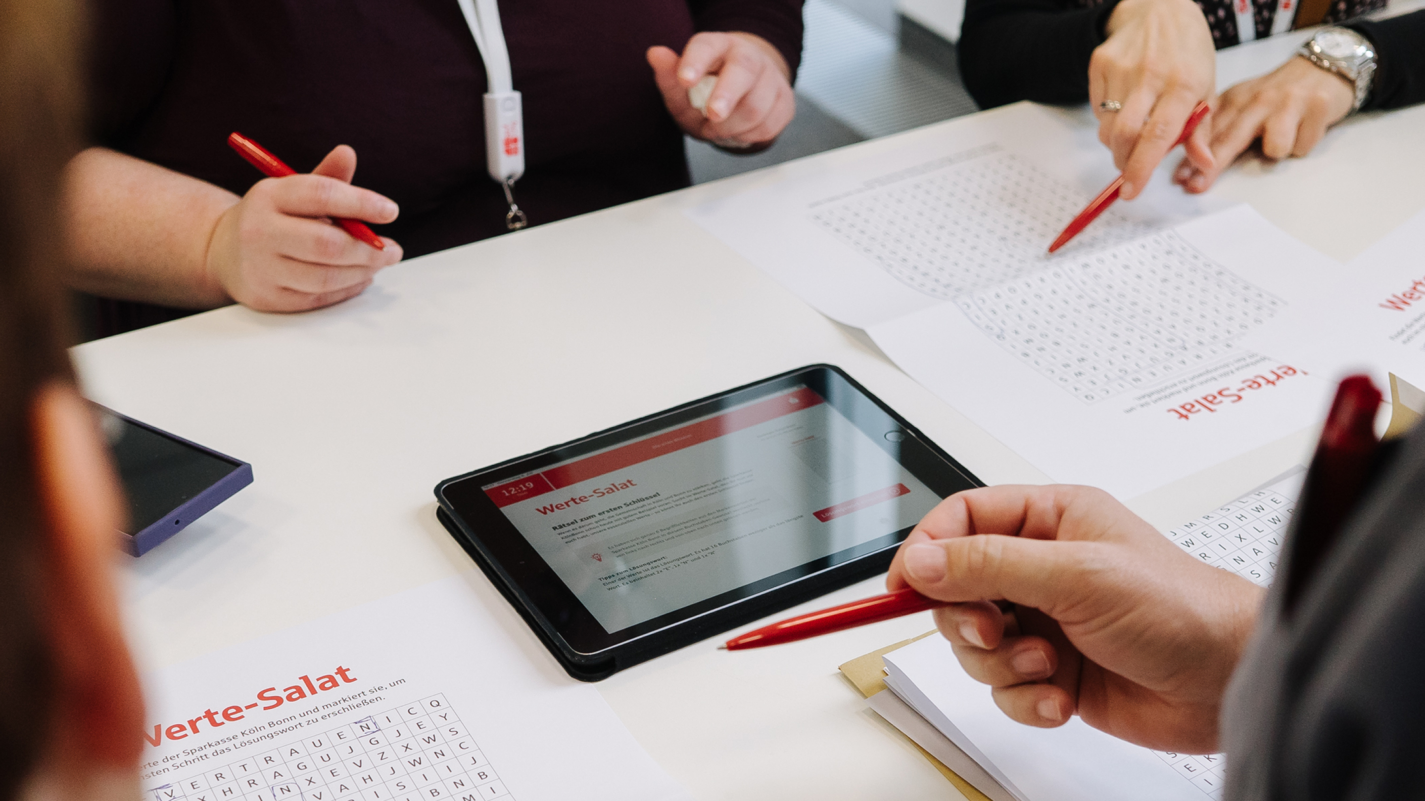 Menschen stehen an einem Tisch um ein Tablet auf dem das Game Changer Business Escape Game geöffnet ist. Von denkwerk