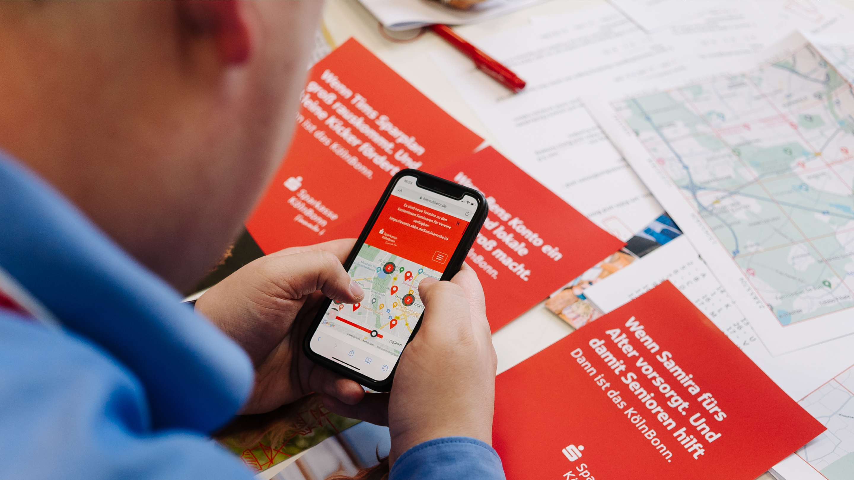 Person mit Handy, im Hintergrund Broschüre Sparkasse KölnBonn