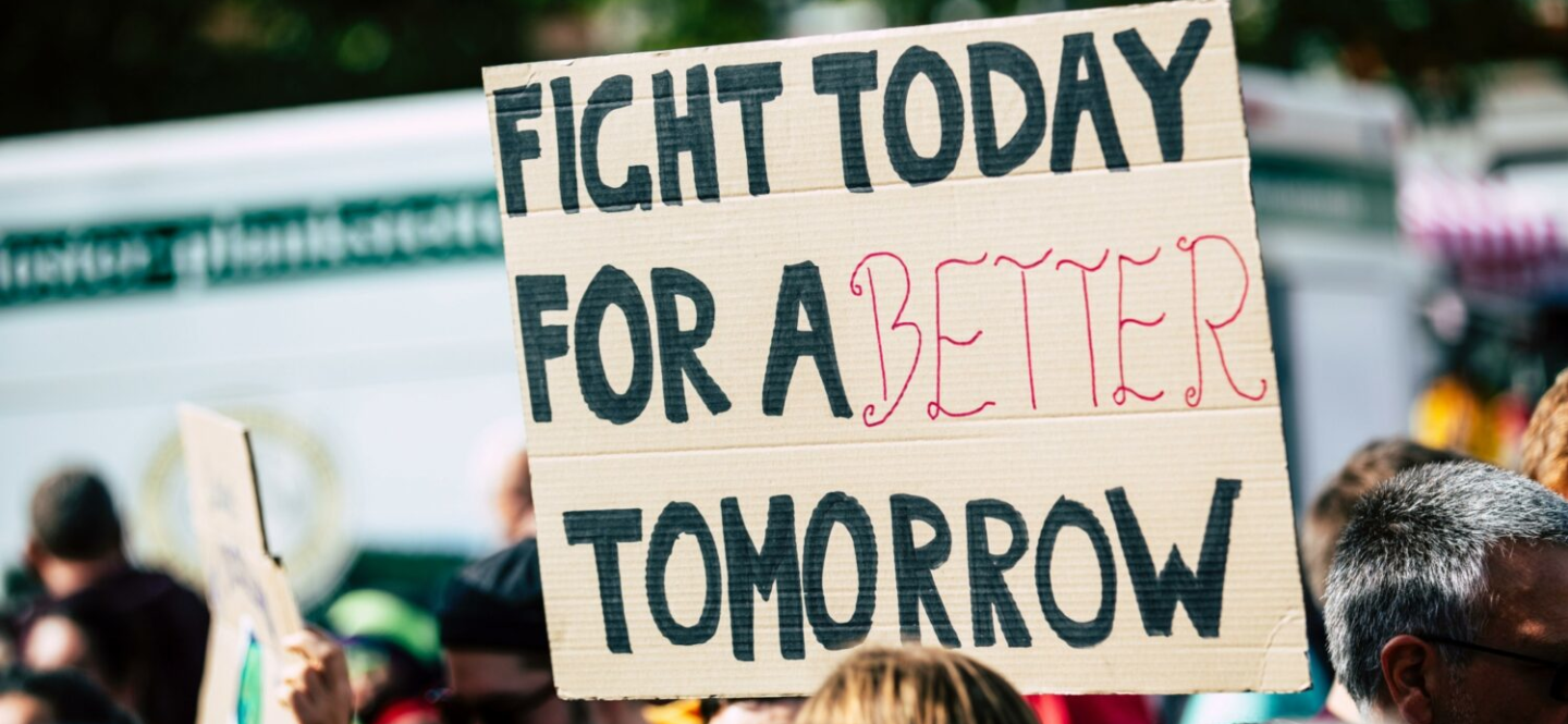 A demonstrator holds a cardboard sign reading "Fight today for a better tomorrow."