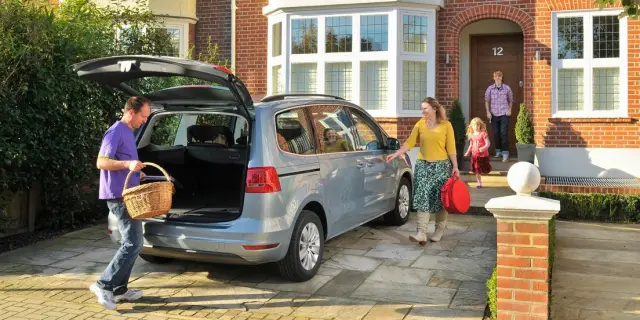 Blue Volkswagen Sharan parked out front of a house, with a family of four getting into the car. The boot is open, with one parent in a purple shirt placing a brown picnic basket in the boot.