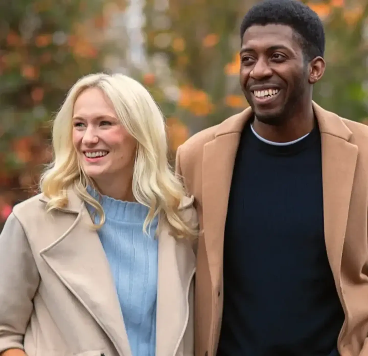 A couple with an arm around each other facing forward, looking away from the camera. Both are smiling and wearing brown coats, with a backdrop of autumnal leaves. Blurred in the background is the front of a red car.
