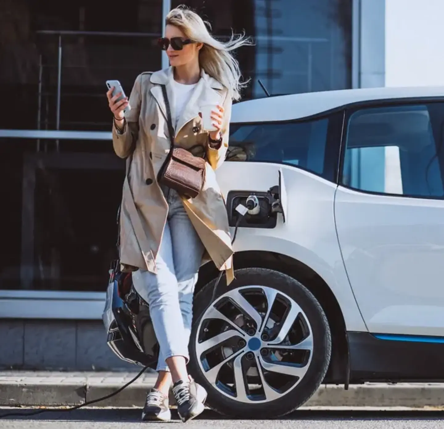 A woman with blonde hair in sunglasses and a tan coat leans against a white electric car that is plugged in and charging.