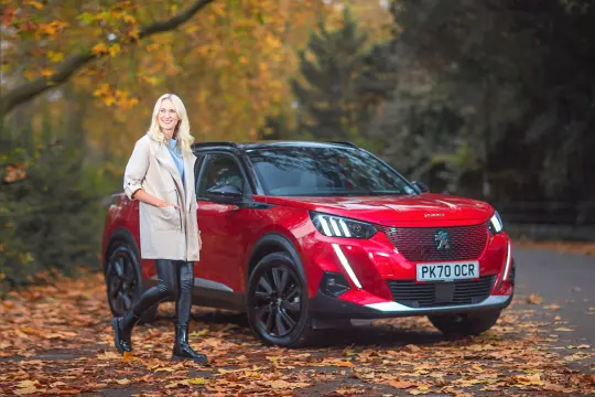 Woman standing next to a red Peugeot e-2008 with an autumnal backdrop