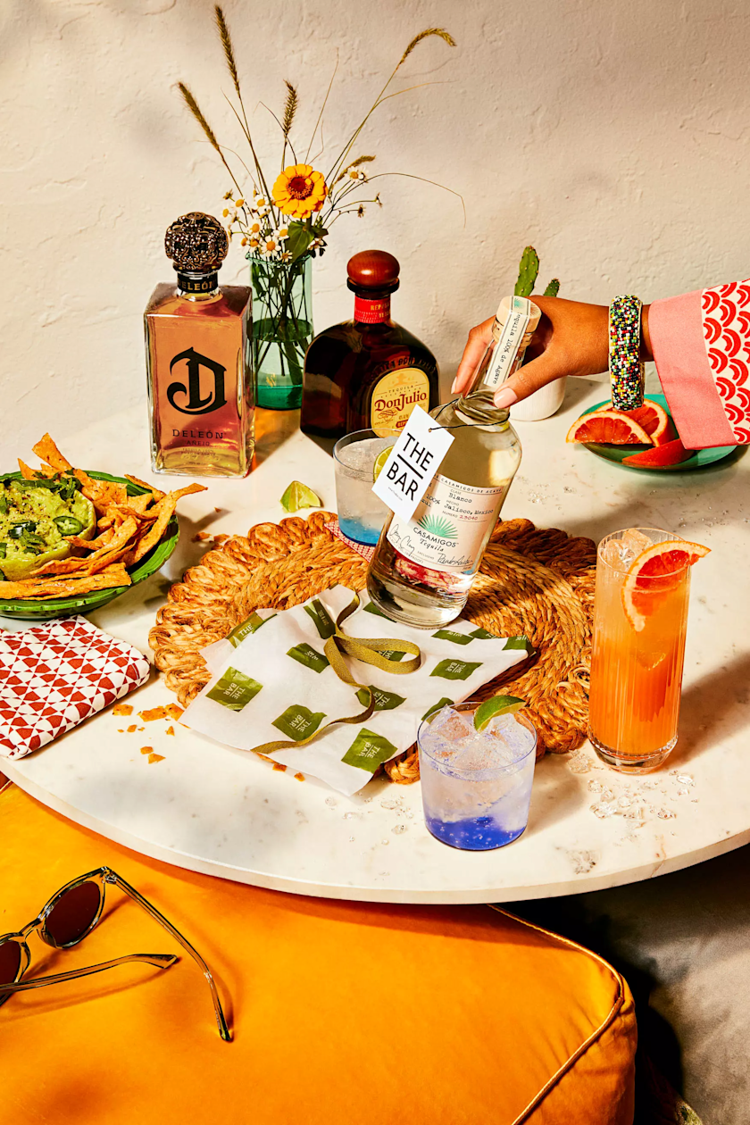 A woman sets down a bottle on a table set for a party with nachos, various mixed drinks, cocktails and bottles of tequila