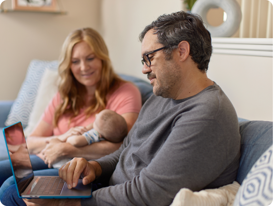 A man and a woman sitting with their newborn baby on a couch in their home. The man is holding and reading on his laptop.