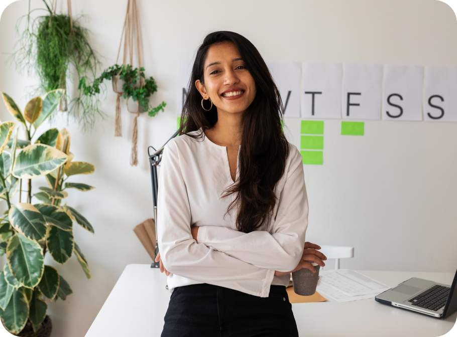 A young woman smiling while situated in a home office setting. Her laptop and paperwork is visible on the desk.