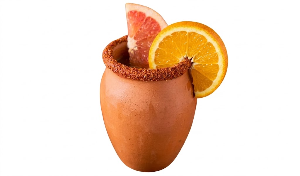 Copo rústico de barro contendo um coquetel. A borda é incrustada com um tempero avermelhado texturizado. A bebida é decorada com uma rodela de laranja na lateral direita e uma fatia de toranja rosada no interior. Fundo branco isolado.