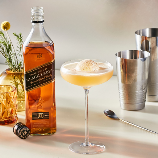 Cóctel en una copa tipo coupé con hielo, rodeado de una botella de Johnnie Walker Black Label Whisky, una coctelera y una cucharilla de bar.
