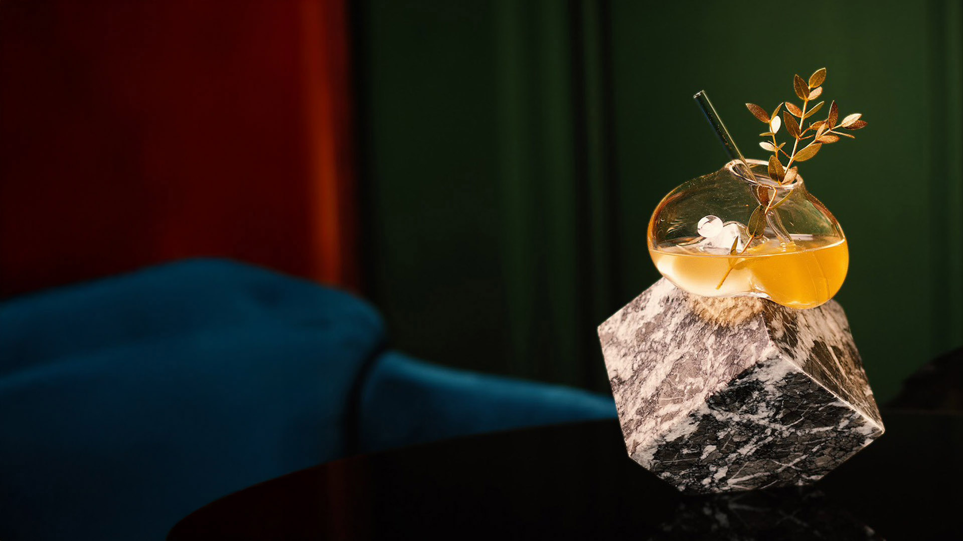 Coquetel dourado em recipiente de vidro orgânico, equilibrado sobre um bloco geométrico de mármore preto e branco. Decorado com um ramo de folhas douradas e canudo de vidro. Fundo elegante e desfocado com poltrona azul de veludo.
