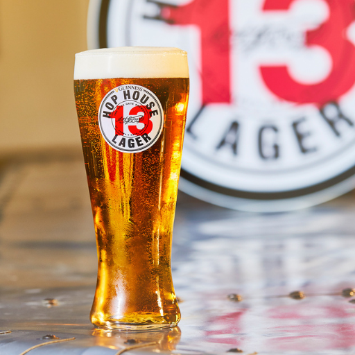 Un vaso de pinta de Hop House 13 Lager en primer plano con un barril de Hop House 13 Lager en el fondo.