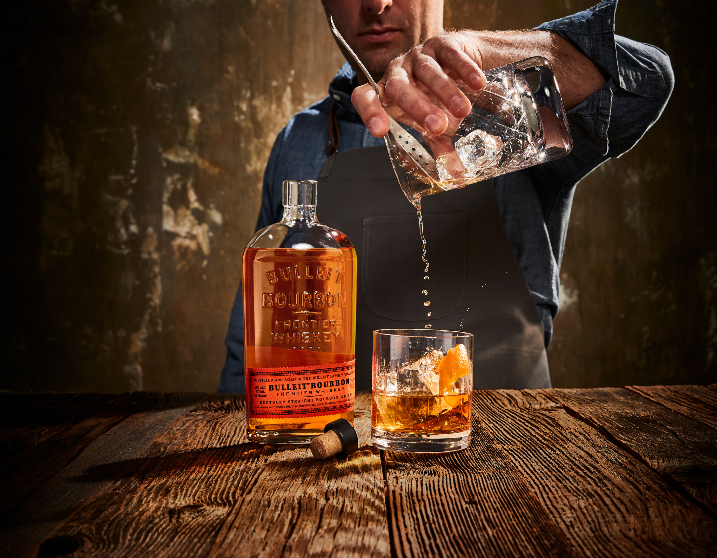 Un hombre cuela un cóctel en un vaso sobre una mesa junto a una botella abierta de whisky Bulleit.