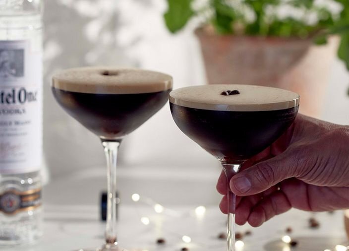 Dos espresso martinis con una botella de Ketel One y luces de colores de fondo. Uno de los martinis está sostenido por una mano en el tallo.