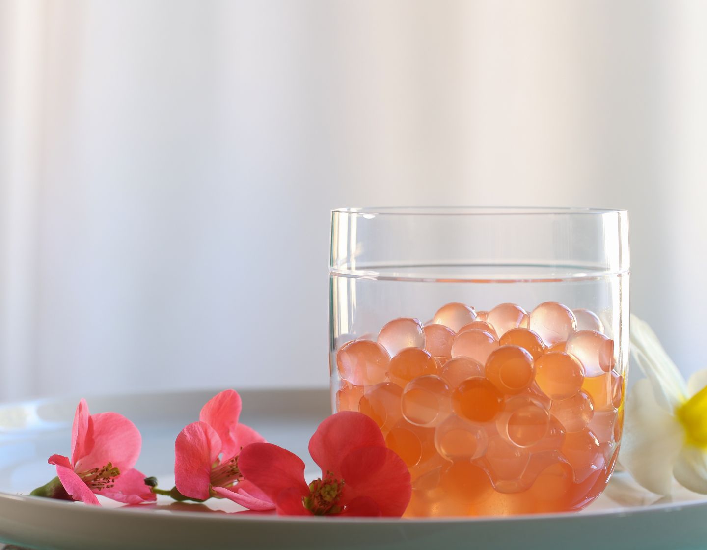 Orange spheres in a short glass next to some tropical flowers on a white plate.