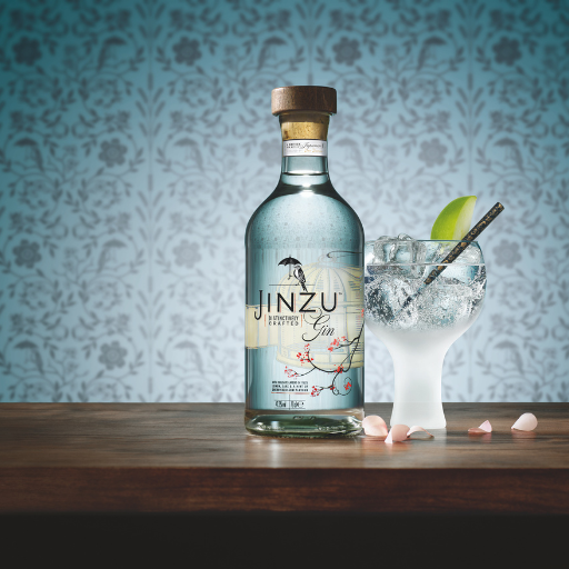 Botella de gin Jinzu en la mesa junto a un vaso de gin con tónica, con un fondo de papel tapiz floral azul.