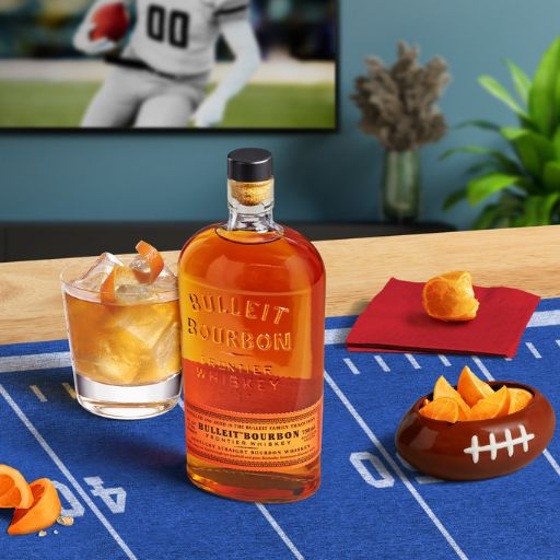 Bulleit_Bourbon_bottle_with_tumblr_glass_of_american_whiskey_and_oranges_on_table_with_sports_tv_in_background
