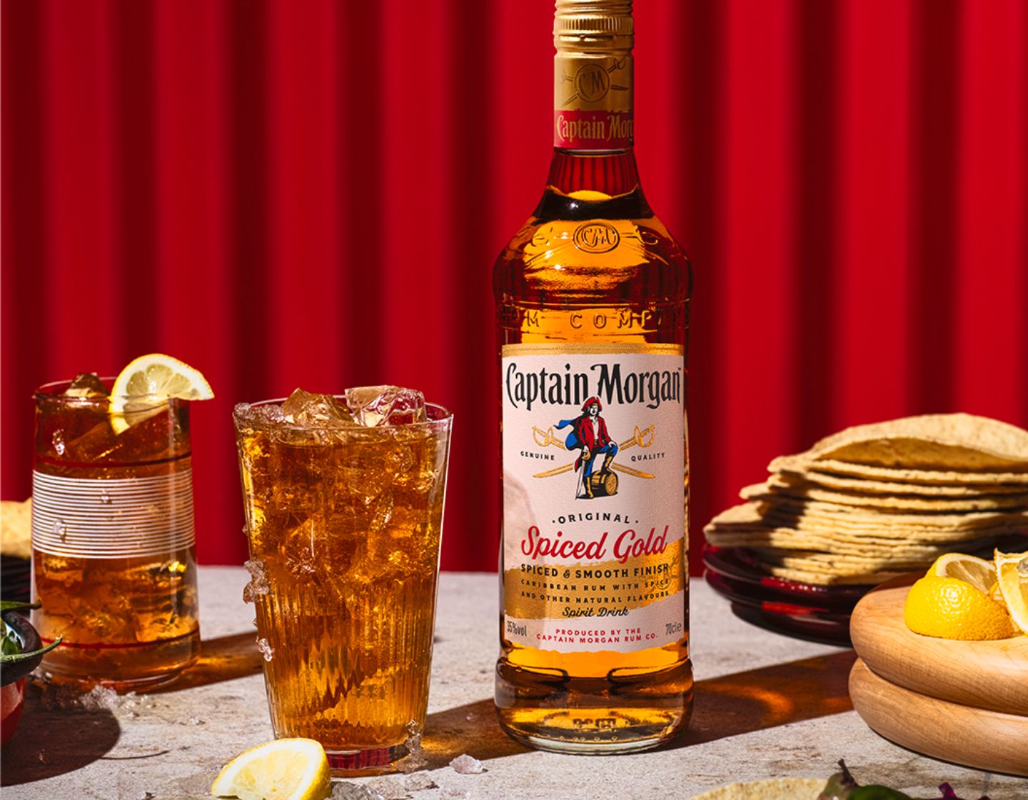 Botella de Captain Morgan Original Spiced Gold sobre una mesa, rodeada de diferentes platos y bebidas, sobre un fondo rojo