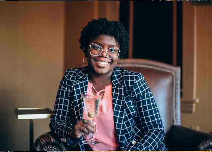 Mixologist and bartender Lauren Paylor sitting in a brown leather chair. She is wearing a checkered suit jacket and a pink blouse. She holds a cocktail and smiles broadly at the camera. 