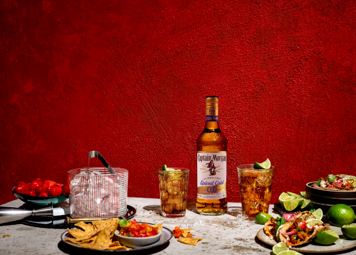 Botella de Captain Morgan Spiced Gold 0.0% Alcohol Free Spirit sobre una mesa rodeada de alimentos y bebidas variadas en un fondo rojo.