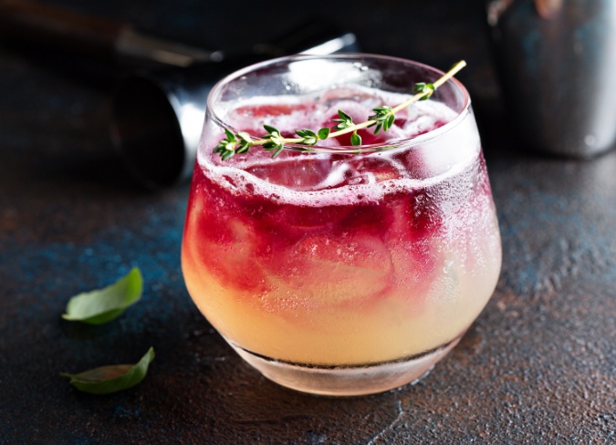 An ombre yellow-pink cocktail with in a short, round glass topped with with an oregano garnish.