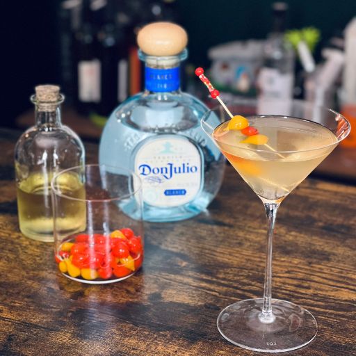 Ein orangefarbener Martini-Cocktail mit einer roten und orangefarbenen Gurkengarnitur und einer Flasche Don Julio Tequila auf einem Holztisch.
