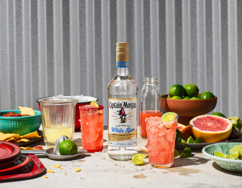 Botella de Captain Morgan White Rum sobre una mesa rodeada de alimentos y bebidas variados, con fondo blanco.