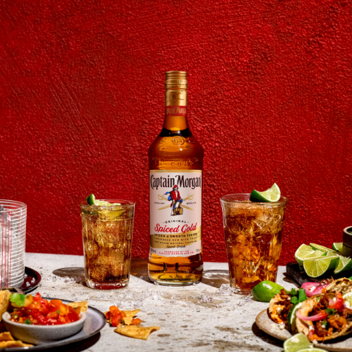 Bottle of Captain Morgan Original Spiced Gold on table surrounded by assorted food and drink against red background