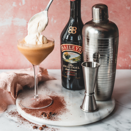 Garrafa de baileys ao lado de um dosador, uma coqueteleira e um drink servido em uma taça e sendo guarnecido com chantilly