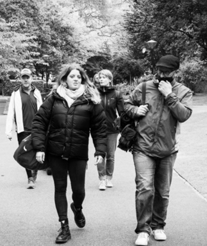 A black and white photo of a group of people. The woman in the front appears to be talking to the man on her side who is adjusting his face mask.    