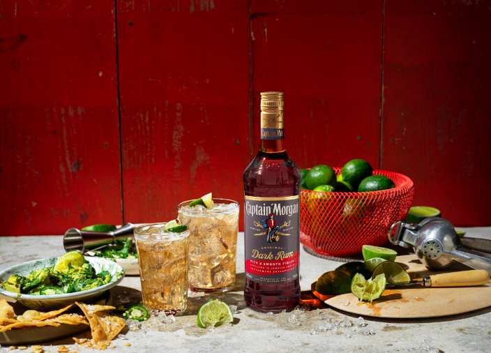 Botella de Captain Morgan Dark Rum sobre una mesa rodeada de comida y bebida variada frente a un fondo rojo.