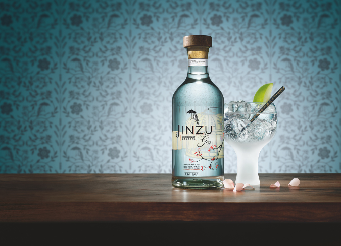 Botella de ginebra Jinzu sobre una mesa junto a un vaso de gin-tonic sobre un fondo de papel pintado con flores azules